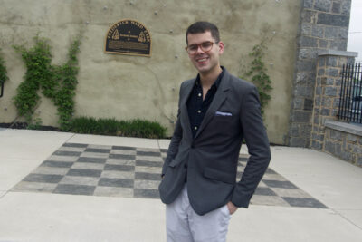 Ryan Nicotra, dressed in a grey suit jacket and light slacks, smiling in front of the stage in the Frederick Ward Armory Park