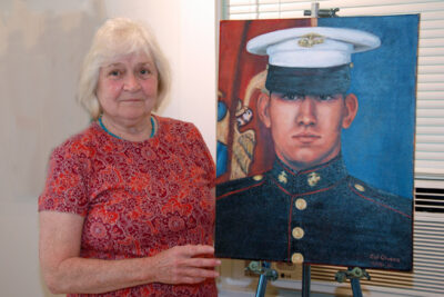 Diae Quick, wearing a red print dress, holds a portait of a veteran on an easel